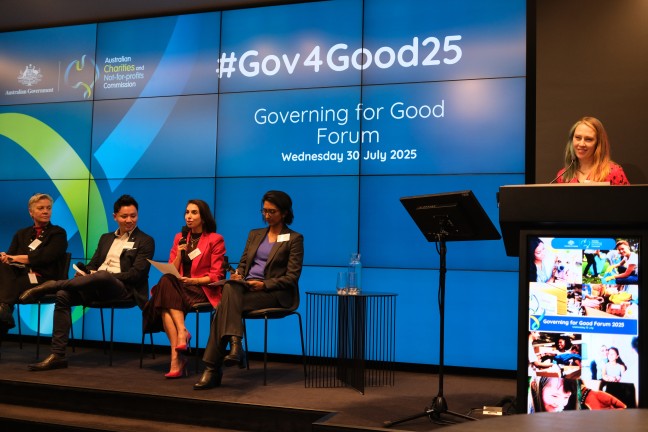 Four people sitting on a stage in front of a digital screen, with a woman to the right standing at a podium.