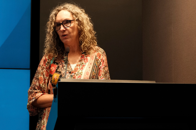 Image of a woman with blonde curly hair standing in front of a microphone and lecturn.